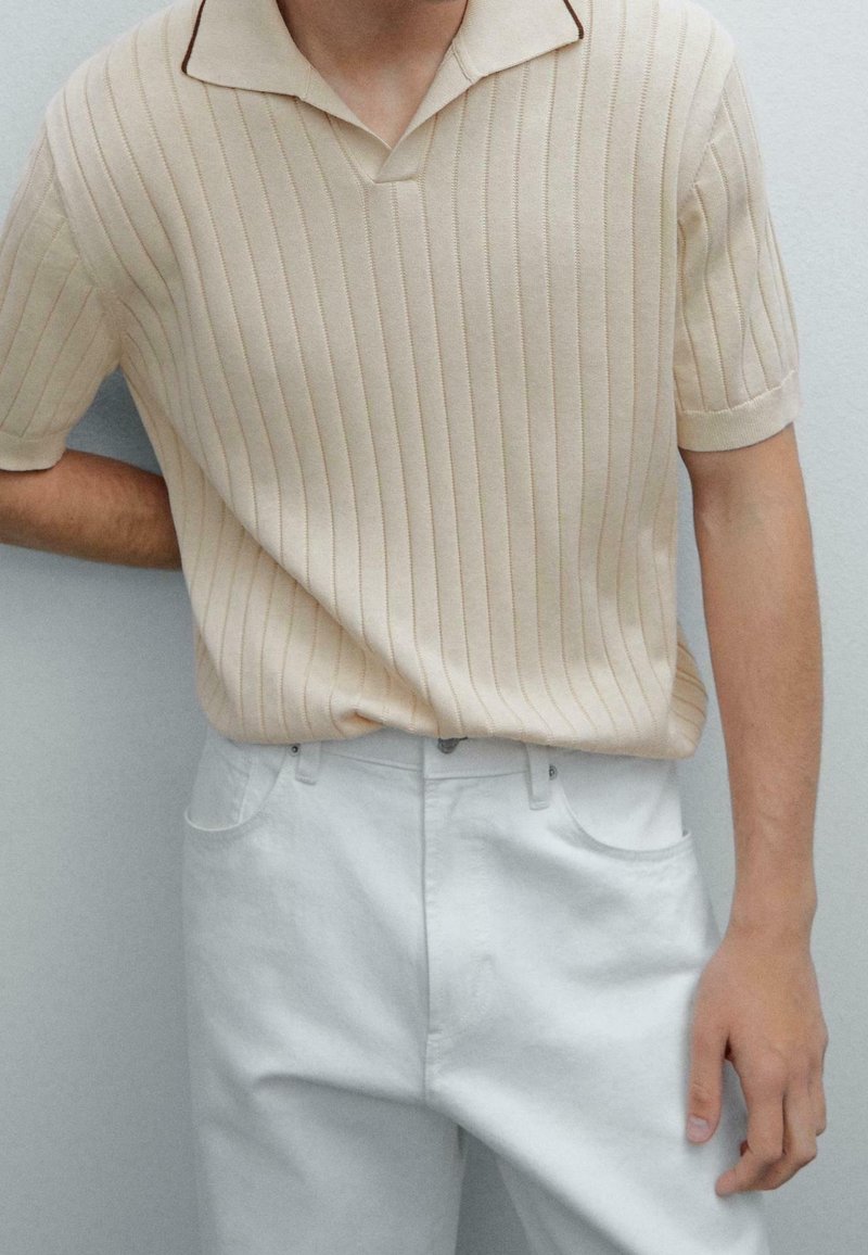 Homme portant un polo beige à manches courtes en maille côtelée, rentré dans un pantalon blanc taille haute, debout contre un mur clair uni.