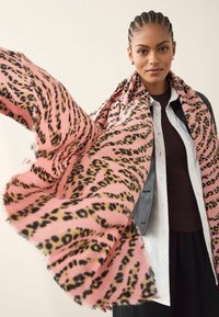 Fular roz cu imprimeu leopard negru, dintr-un material moale, cu formă dreptunghiulară, marginile franjurate, ținut de o persoană care poartă o jachetă de piele neagră.