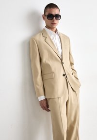 Beige jakkesæt lavet af tekstureret stof, med en enkeltknapsjakke, spidse klippet lapeller og sorte knapper, parret med en hvid skjorte.