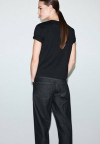 Crna majica s kratkim rukavima i preklopljenim rukavima, u kombinaciji s tamnim trapericama. Vidljiva glatka tekstura tkanine i klasičan kroj sa stražnje strane.