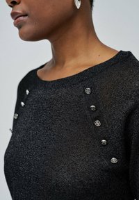 Nahaufnahme einer Person, die einen schwarzen strukturierten Pullover mit dekorativen silbernen Knöpfen an der Schulter und silbernen Creolen trägt.