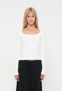 Blonde vrouw in een witte langärmige top met ronde hals en losse zwarte broek met trekkoord, staand tegen een effen witte achtergrond.