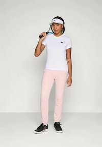 Hvit kortermet skjorte med en liten svart logo, parret med lyserosa joggebukser og svarte joggesko, og holder en tennisracket.