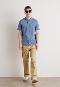 Cămașă polo din bumbac de culoare bleu deschis, cu mâneci scurte, pantaloni din bumbac bej și adidași albi. Modelul poartă ochelari de soare negri.