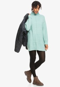 Mintgrön oversized stickad tröja med hög krage, kombinerad med svarta leggings och bruna ankboots. Modellen håller en svart jacka.