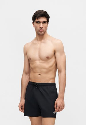 Mężczyzna bez koszulki z krótkimi ciemnymi włosami, ubrany w czarne kąpielówki z białym logo, stoi na tle jednolitego jasnego tła.