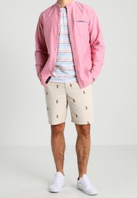 Chemise à manches longues rose avec des boutons sur un T-shirt rayé, associée à un short crème agrémenté de petits motifs floraux verts et rouges, et des baskets blanches.