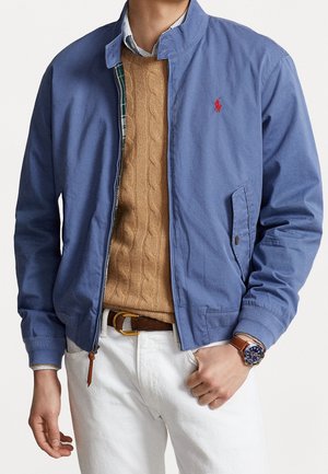 Homme portant une veste zippée bleue sur un pull en maille torsadée beige, pantalon blanc, ceinture marron et montre en cuir marron au poignet gauche.