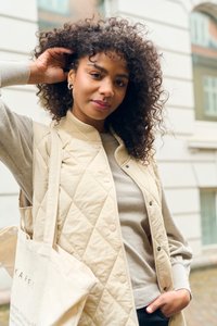 Gilet matelassé beige avec un col côtelé sur un pull gris clair. Le mannequin tient un sac fourre-tout en toile, affichant des cheveux bouclés et une expression naturelle.