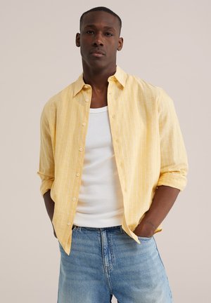 Man draagt een geel gestreept overhemd over een wit hemd, met handen in de zakken van lichtblauwe spijkerbroek, staand tegen een effen achtergrond.