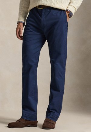 Mann trägt marineblaue Hose, braune Wildleder-Loafers, cremefarbenen Kabelstrickpullover und eine Uhr am linken Handgelenk.