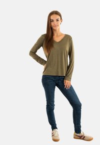 Top verde oliva a maniche lunghe con scollo a V e dettagli in bottoni, abbinato a jeans skinny blu scuro e sneakers chiare con strisce bordeaux.