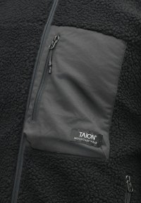 Giacca in pile nera con parte superiore morbida e testurizzata e tasca sul petto nera e liscia, dotata di chiusura con zip e etichetta sull'orlo.