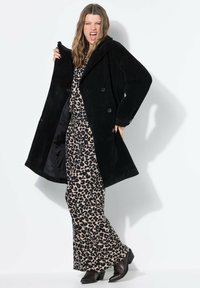 Žena s dvojím zeleným líčením očí, oblečená v černém kabátu přes leopardí šaty a černé boty, pózuje s otevřenými ústy proti bílé zdi.