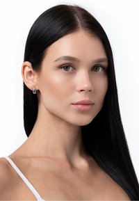 Srebrni uhani v obliki kapljice, pritrjeni na ušesu modela, z sijočim zaključkom in poenostavljenim dizajnom. Lasje so ravni in črni. Ozadje je belo.