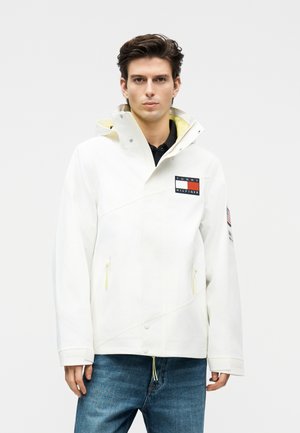 Giovane uomo che indossa una giacca bianca Tommy Hilfiger con toppe con il logo, tasche con zip e colletto alto, abbinata a jeans blu.