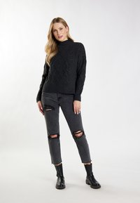 Sort kabelstrikket sweater med høj hals, parret med grå slidte jeans med revner ved knæene, og sorte ankelstøvler.