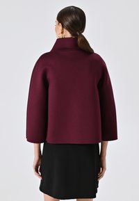 Burgunderfarbenes Poncho-Oberteil mit hohem Kragen, 3/4-Ärmeln und glatter Textur, kombiniert mit einem schwarzen, figurbetonten Rock.