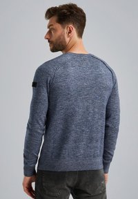 Blauer Strickpullover mit Raglanärmeln, strukturiertem Stoff und einem kleinen Logo-Patch am Ärmel. Sichtbare gerippte Bündchen und Saum.