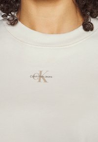 Felpa beige chiaro con colletto a girocollo a costine. Presenta un piccolo logo "CK" in rilievo e la scritta "Calvin Klein Jeans" in un sottile colore dorato.