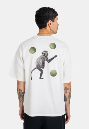 Man with tattoos wearing white t-shirt featuring a graphic of a monkey juggling three green balls on the back.