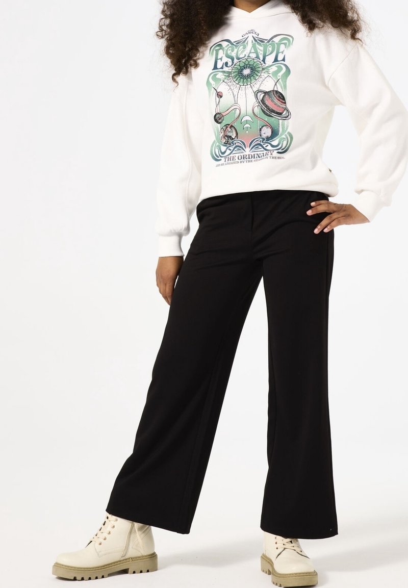 Personne portant un sweat à capuche blanc avec un motif "Escape" sur le thème de l'espace en vert, un pantalon noir à jambes larges et des bottines lacées crème.