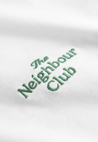 Texte vert brodé "The Neighbour Club" sur tissu blanc, montrant la texture du tissu et les détails de la couture.