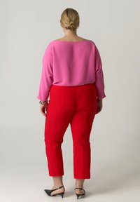 Fuchsia blouse met een boothals en lange mouwen, gecombineerd met rode aangesloten broeken en zwarte pumps met puntige neus. Subtiele textuur in de stof.