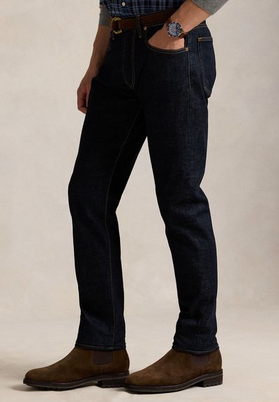 Férfi sötétkék farmert, barna velúr Chelsea csizmát, barna övet, kék kockás inget, szürke pulóvert és barna szíjas karórát visel.