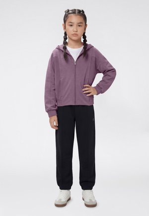 Junges Mädchen mit geflochtenem Haar, trägt einen lila Kapuzenpullover mit Reißverschluss, schwarze Hose, weiße Turnschuhe, steht mit einer Hand in der Hüfte.