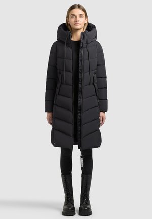 Femme portant un long manteau matelassé noir avec capuche, un pantalon noir et des bottes noires à lacets, debout devant un fond uni.