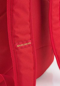Mochila roja hecha de un tejido duradero, con una textura suave, costuras reforzadas y una etiqueta con el logotipo dorado "SKECHERS" en el lateral.