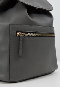 Sac à dos en cuir gris avec rabat pliant, poche zippée ton or et texture lisse. Comprend une étiquette en cuir amovible.