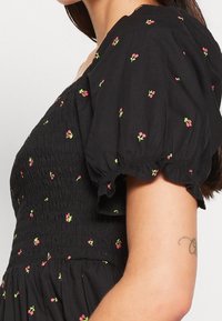 Robe noire avec un corsage smocké et des manches bouffantes, ornée de petites broderies florales rouges et vertes, et une jupe fluide.