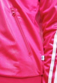 Rosa träningsjacka med ficka som stängs med dragkedja, slät tyg, ribbad fåll och vita sidosträck med en blå logotyp på fållen.