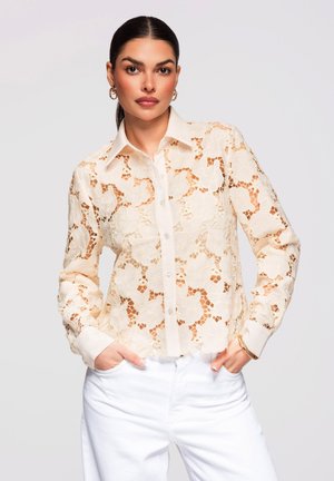 Femme aux cheveux foncés portant une chemise boutonnée en dentelle florale beige transparente et un pantalon blanc, debout les mains dans les poches devant un fond uni.