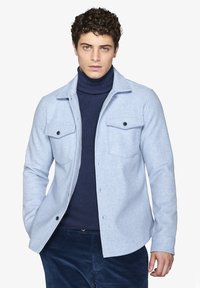 Lyseblå skjorte med knapper og to brystlommer, lavet af et struktureret stof, båret over en marineblå ribbet rullekravesweater.