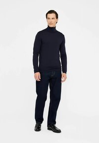 Námořnický rolák z měkkého materiálu, kombinovaný s tmavě modrými džínami. Model stojí, ruce podél těla, obutý v černých botách.