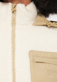 Chaqueta de forro polar blanca con cremallera beige, cuello alto y un bolsillo beige acolchado con detalle de costura. Textura suave en todo el conjunto.