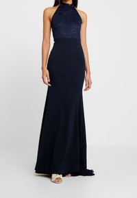Granatowa wieczorowa suknia z koronkowym dekoltem na szyi i dopasowanym dołem. Miękka tkanina opada na podłogę, uzupełniona neutralnymi butami.