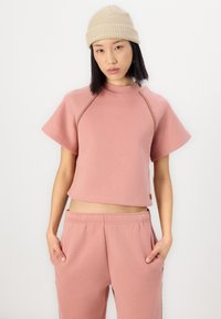 Kurzärmliger, verkürzter pinker Sweatshirt mit weißen Nähten. Kombiniert mit passenden pinken Hosen mit seitlichen Taschen. Mütze in hellbeige.