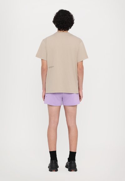 Beige bavlněné tričko s krátkým rukávem, kombinované se světle fialovými šortkami. Černé boty a ponožky doplňují celý outfit. Textová detail na tričku.