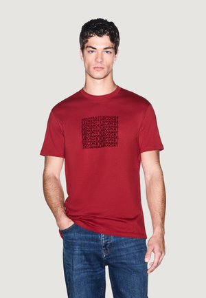 Młody mężczyzna z krótkimi kręconymi włosami nosi czerwony t-shirt Armani Exchange z czarnym logo i niebieskie dżinsy, stoi na jednolitym tle.