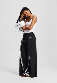 Zwarte wijde broek met witte zijknopen en bies, gecombineerd met een witte tanktop. Accessoires zijn onder andere een witte tas met zwarte letters.
