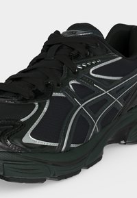 Chaussure de sport noire avec des accents argentés, semelle texturée et lacets noirs, conçue pour la course ou l’usage sportif.