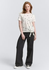 Biała koszulka z krótkim rękawem w czarne i różowe serca, z węzłem w talii. W połączeniu z luźnymi czarnymi dżinsami i trampkami.