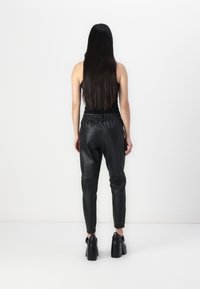 Vrouw met lang donker haar, gekleed in een mouwloos zwart topje, zwarte leren broek en zwarte platformhakken, staand tegen een effen witte achtergrond.