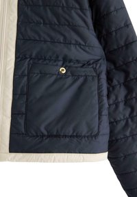 Mørkeblå pufjakke med cremefarvede detaljer, der har en horisontal quiltet tekstur, lynlås lukning og en frontlomme med en guld trykknap.