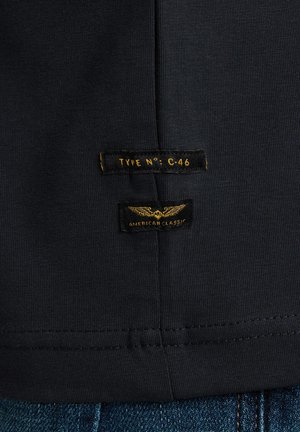 Schwarzes Baumwoll-T-Shirt mit glatter Textur. Verfügt über zwei Etiketten: "TYPE NO: C-46" und "AMERICAN CLASSIC" in Gelb auf dunklem Stoff.
