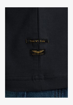 T-shirt en coton noir avec une texture lisse. Il comporte deux étiquettes : "TYPE NO: C-46" et "CLASSIQUE AMÉRICAIN" en jaune sur un tissu sombre.